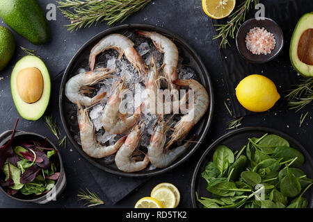 Freschi gamberoni in cucina con insalate, erbe, limoni e avocado su pietra nera tavolo, vista dall'alto Foto Stock