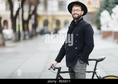 Fiducioso e elegante. Lunghezza completa di fiducioso giovane uomo barbuto regolazione eyewear e guardando la fotocamera mentre è seduto sulla sua bicicletta all'aperto Foto Stock