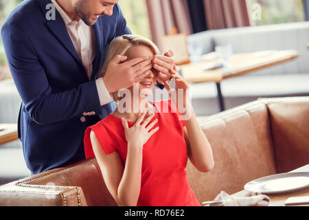 Una giovane coppia in data nel ristorante uomo in piedi dietro la donna seduta che copre i suoi occhi rendendo felice sorpresa Foto Stock