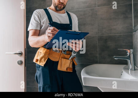 Vista parziale dei maschi di idraulico con toolbelt iscritto negli appunti vicino al lavandino rotti nella stanza da bagno Foto Stock