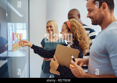 Sorridente giovane imprenditrice la lettura di foglietti adesivi su una parete di vetro mentre il brainstorming con i colleghi in un ufficio Foto Stock