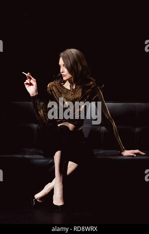 Donna glamour seduta sul lettino e il fumo di sigaretta su isolato nero Foto Stock