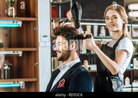 Bella giovane parrucchiere sorridente in telecamera durante l'asciugatura e pettinatura dei capelli belli al client in un salone di bellezza Foto Stock