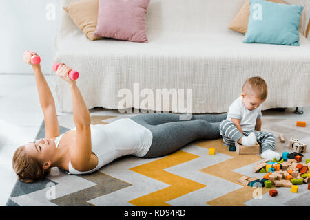 Donna attraente fare esercizio con manubri e simpatico bimbo ragazzo giocando con multicolore di cubi di legno sul tappeto in salotto Foto Stock