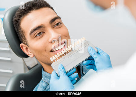 Close up bello african american uomo sorridente vicino a denti tavolozza di colori prima della procedura di sbiancamento Foto Stock