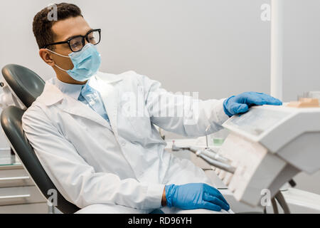 African American dentista in seduta occhiali, maschera e mantello bianco Foto Stock