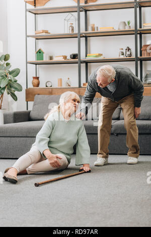 Senior moglie seduta sul pavimento con dolore in ginocchio vicino al marito in pensione Foto Stock