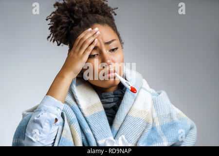Donna malata con il termometro nella sua bocca, avvolti in plaid. Foto di African American donna avvolta in pagato su sfondo grigio. Il concetto di assistenza sanitaria. Foto Stock