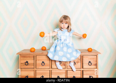 Bambino Toddler Baby girl in azzurro vestito seriamente seduto sulla cassa di legno dei cassetti e azienda arancio freschi Mandarini nelle sue mani sul rombo di sfondo. Bambini sentimenti emozioni Foto Stock