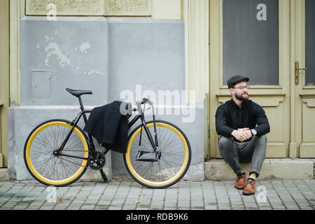 Giovane bello hipster uomo seduto sulla strada e in posa con zaino e bicicletta sul tramonto Foto Stock