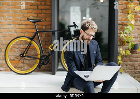 Attraente giovane uomo barbuto lavora con il computer portatile seduto sul pavimento in strada con la bicicletta Foto Stock