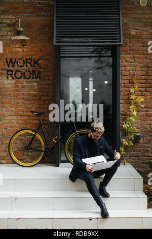 Ritratto di giovane bello uomo barbuto che cercano riposo utilizzando un computer portatile con bicicletta all'esterno. Foto Stock