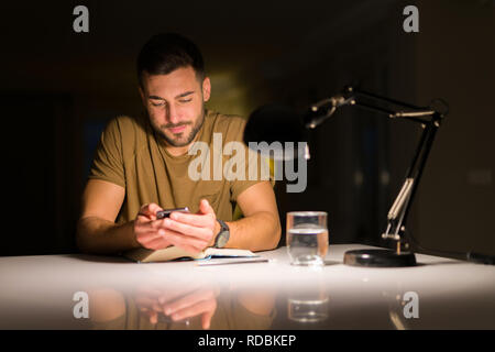 Giovane uomo bello studiare a casa , utilizzando lo smartphone, guardando il telefono sorridente Foto Stock