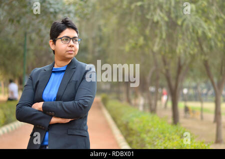 Closeup ritratto di un fiducioso giovani indiani professionista aziendale di donna con capelli corti e occhiali, attraversato mani piegate in un ambiente esterno Foto Stock