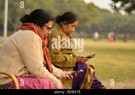 Nord e sud indiane senior donne anziane utilizzando la tecnologia occupato sul loro smartphone in un parco all'aperto a Delhi inverni. Foto Stock