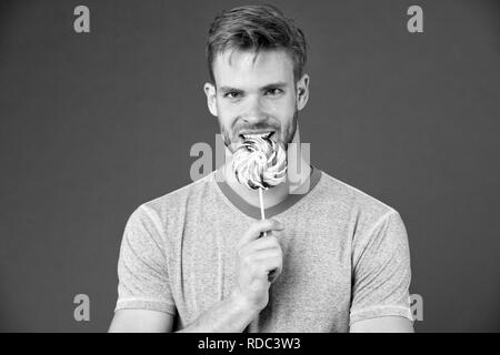 Chi si preoccupa di dieta. L'uomo morde grande sweet lollipop. Uomo con setola ama i dolci. Dieta viziati. Atleta sulla faccia sorridente mangia lecca-lecca colorati, sfondo violetto. Cheat concetto di pasto. Foto Stock