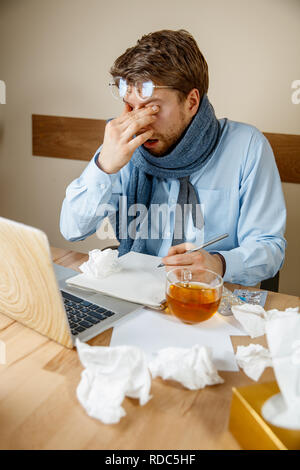 La sensazione di malessere e stanco. Frustrato triste malati infelice giovane massaggiare i suoi occhi mentre è seduto al suo posto di lavoro in ufficio. L'influenza stagionale, una pandemia di influenza, prevenzione di malattia concetto Foto Stock