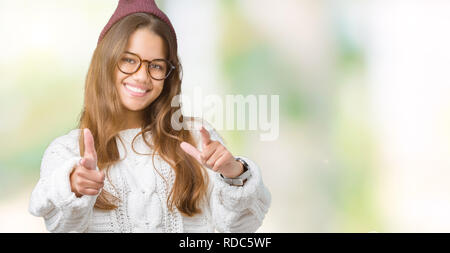 Giovane bella bruna hipster donna che indossa occhiali e cappello invernale su sfondo isolato dita di puntamento per fotocamera con felice e divertente volto. G Foto Stock