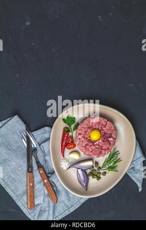 Deliziosa la tartare di manzo con tuorlo d'uovo e gli ingredienti sulla piastra in ceramica, set di posate coltello, forchetta sulla pietra nera di calcestruzzo superficie testurizzata sfondo. Co Foto Stock