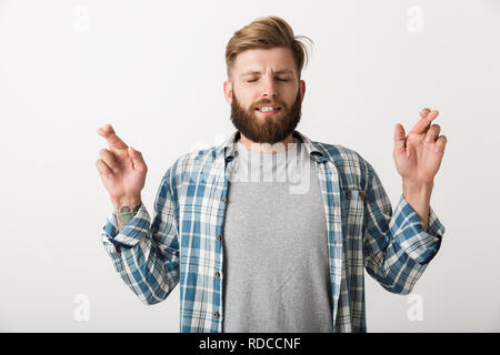 Preoccupato uomo barbuto vestito in plaid shirt in piedi isolato su sfondo bianco, tenendo le dita incrociate per buona fortuna Foto Stock