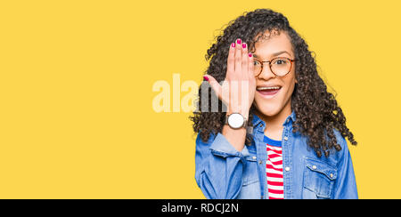 Giovane bella donna con capelli ricci con gli occhiali che coprono un occhio con la mano con fiduciosa sorriso sul volto e la sorpresa di emozione. Foto Stock