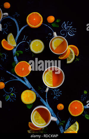 Tè al limone chalk tree con teacups, arance e le fette di limone e foglie di menta e kumquat. Bevanda calda piatti creativi giacciono su uno sfondo scuro con spazio di copia Foto Stock