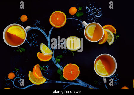 Tè al limone chalk tree con teacups, arance e le fette di limone e foglie di menta e kumquat. Bevanda calda piatti creativi giacciono su uno sfondo scuro con spazio di copia Foto Stock