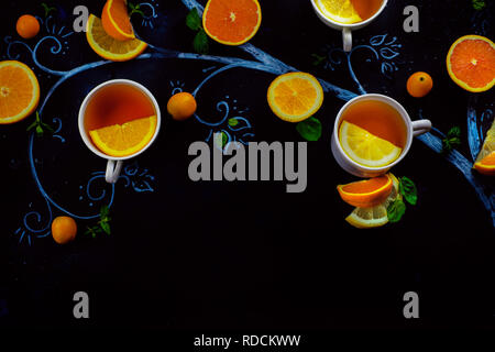 Tè al limone chalk tree con teacups, arance e le fette di limone e foglie di menta e kumquat. Bevanda calda piatti creativi giacciono su uno sfondo scuro con spazio di copia Foto Stock