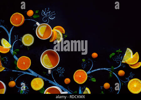 Tè al limone chalk tree con teacups, arance e le fette di limone e foglie di menta e kumquat. Bevanda calda piatti creativi giacciono su uno sfondo scuro con spazio di copia Foto Stock