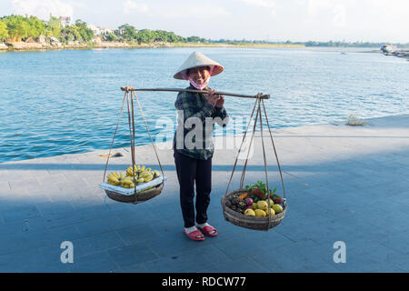 Donna vietnamita che trasportano cesti di frutta su una spalla polo o milkmaid la forcella nella città vietnamita di Hoi An in Quang Nam Provincia Foto Stock