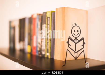 Libri su uno scaffale di legno con un bastone figura su un libro aperto seduta holding e la lettura di un libro e sorridente Foto Stock