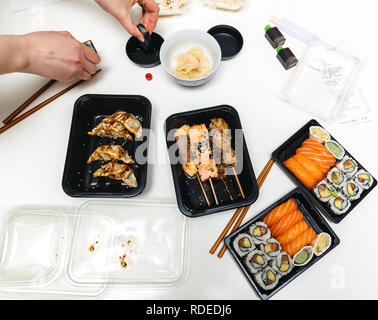 Tabella contenente il sushi casella consegna con sushi california, Maki, sashimi, yakitori e bastoni di legno con wasabi, gyoza e salsa di soia - donna preparare la salsa di soia Foto Stock