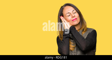 Bella la mezza età donna che indossa Gilet invernale sonno stanco di sognare e in posa con le mani insieme mentre sorridente con gli occhi chiusi. Foto Stock