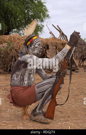 Karo guerriero con del corpo e del viso dipinti e un fucile seduto sul suo appoggiatesta, Omo river valley, l'Etiopia meridionale, Africa Foto Stock