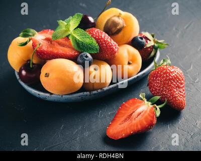 Vista ingrandita di frutti e bacche su sfondo scuro. Cumulo di fragole fresche, mirtilli, albicocca e foglie di menta nella piastra alla moda. Focus su fragola. Cibo sano,super,la dieta,detox concetto. Foto Stock