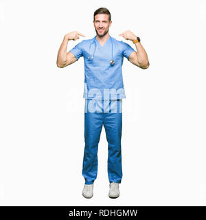 Bello medico uomo che indossa medical uniforme su sfondo isolato guardando fiduciosi con il sorriso sul volto, rivolto a se stessi con le dita in fieri e hap Foto Stock