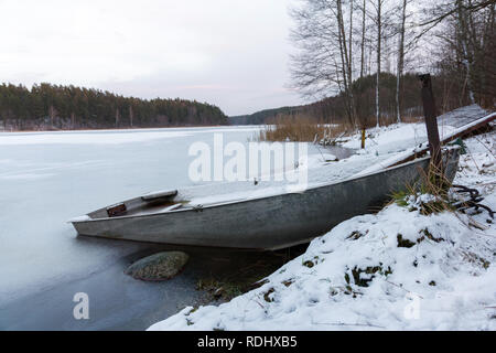 Barca congelate in ghiaccio sul lago di costa con alberi e foresta in background in inverno Foto Stock