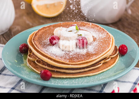 Pila di frittelle ricoperte di miele, zucchero a velo, banana e mirtilli rossi. Su uno sfondo di legno in una piastra di colore blu Foto Stock