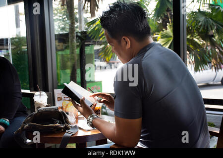 Thai uomo seduto rilassato libro di lettura e parlare di bere caffè ghiacciato in cafe shop on November 13, 2018 in Saraburi, Thailandia Foto Stock