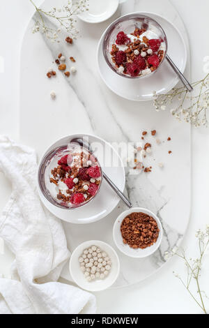 Laici piana con due bicchieri di più livelli di cereali e yogurt con lamponi su un vassoio in marmo Foto Stock