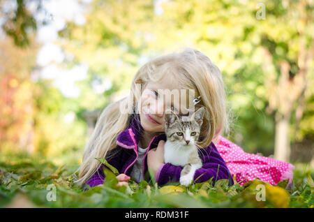 Bella ragazza bionda con cat e giacente insieme sull'erba nel periodo autunnale Foto Stock