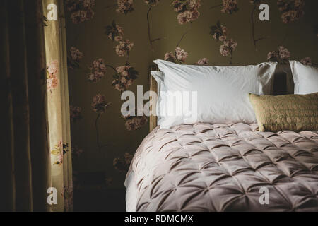 Vista interna della camera da letto con tende e carta da parati con motivo floreale, rosa pallido piumini e cuscini bianchi sul letto matrimoniale. Foto Stock