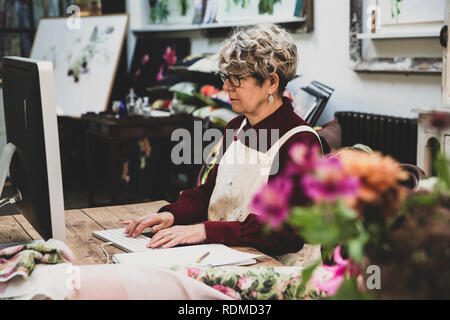 Senior donna che indossa gli occhiali, vestito rosso e bianco grembiule seduti al tavolo di legno in un studio, lavorando sul computer desktop. Foto Stock