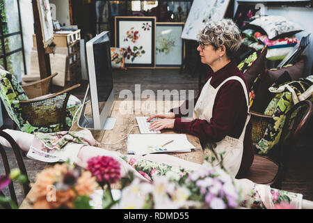 Senior donna che indossa gli occhiali, vestito rosso e bianco grembiule seduti al tavolo di legno in un studio, lavorando sul computer desktop. Foto Stock