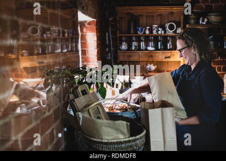 Donna sorridente con gli occhiali immissione appena sfornato torte in carta marrone shopping bag. Foto Stock