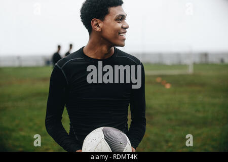 Sorridente giocatore di calcio in piedi sul campo tenendo un calcio che guarda lontano. Allegro giocatore di calcio la pratica al mattino sul campo con i compagni di squadra giocare Foto Stock