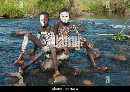 Due ragazzi Surma con trattamenti per il viso e per il corpo dipinto seduto su una roccia nel fiume, Kibish, Omo River Valley, Etiopia, Africa Foto Stock