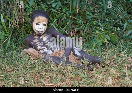 Surma ragazzo con trattamenti per il viso e per il corpo dipinto giacente su erba, Kibish, Omo River Valley, Etiopia, Africa Foto Stock