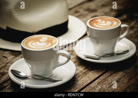 Due tazze di cappuccino/ cappuccino con schiuma arte su una tavola di legno tavolo rustico con un cappello Fedora in background. Rilassatevi, relax, brunch, poggiando su una bre Foto Stock