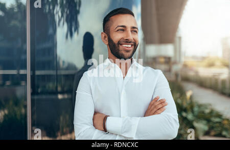 Ritratto di un giovane fiducioso sorridente uomo indiano con le braccia incrociate guardando in lontananza Foto Stock
